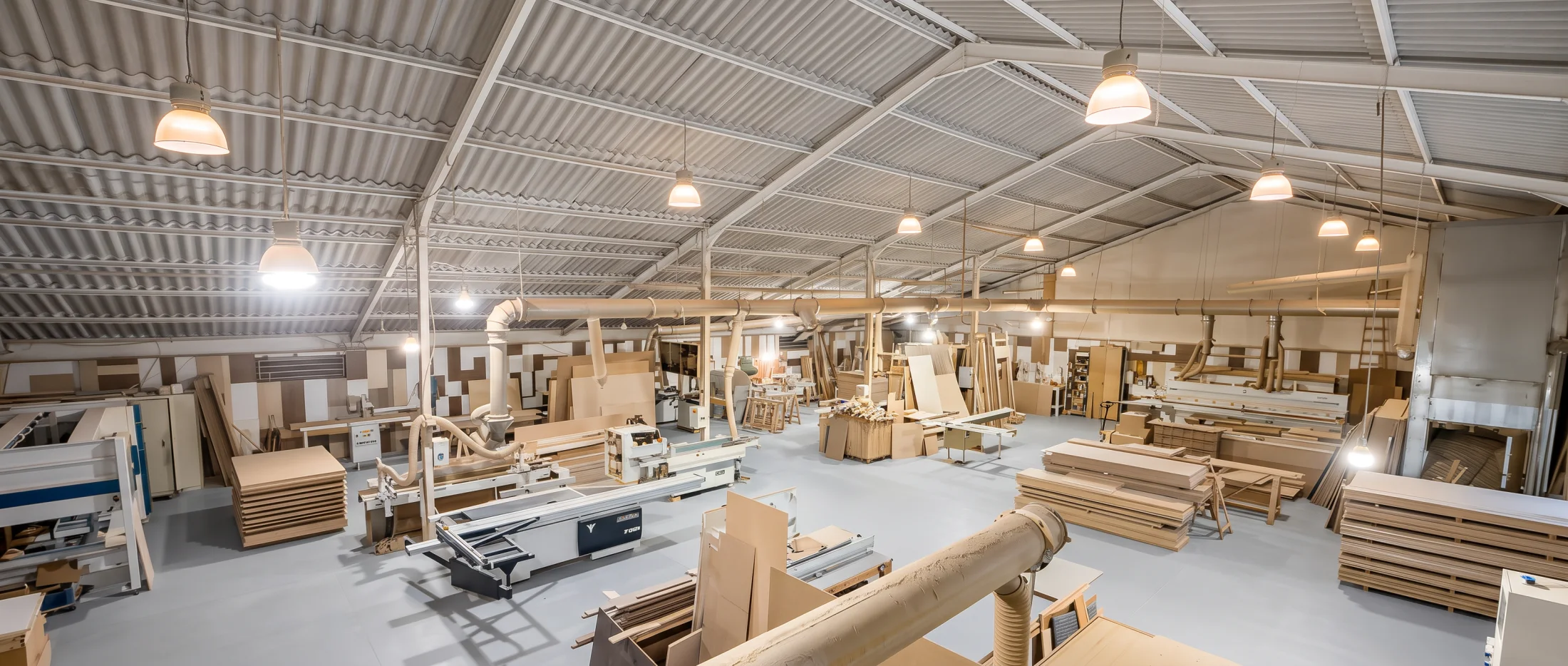 La halle de l'atelier vue d'en haut : toit ondulé sur fermes blanches, scie à panneaux, tubes d'aspiration et piles de panneaux rangées.